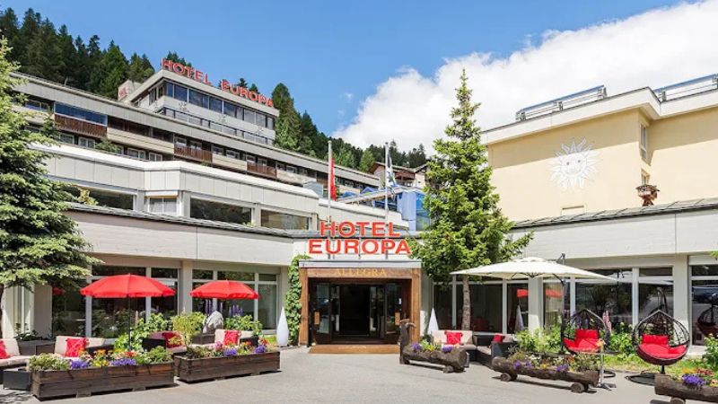 Entr� till Hotel Europa med modern fasad, r�da parasoller och sittgrupper i en blomsterprydd uteplats omgiven av gr�na tr�d och berg.
